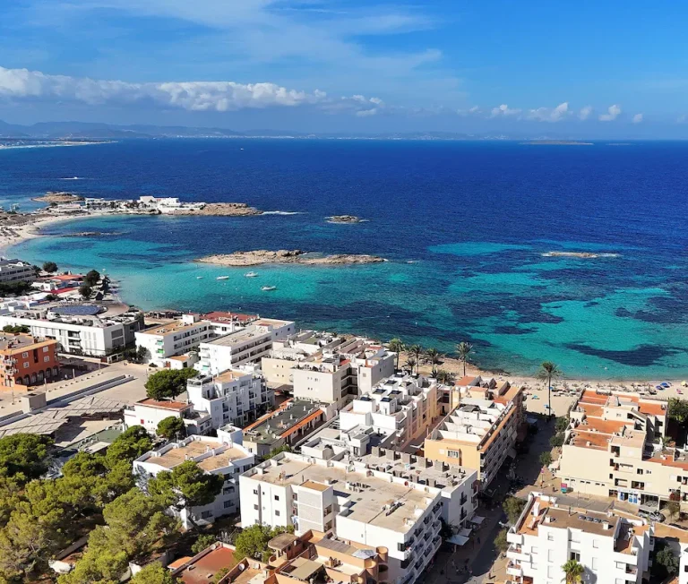 Formentera Vue panoramique d'une plage et de la mer turquoise, avec des bâtiments en arrière-plan.